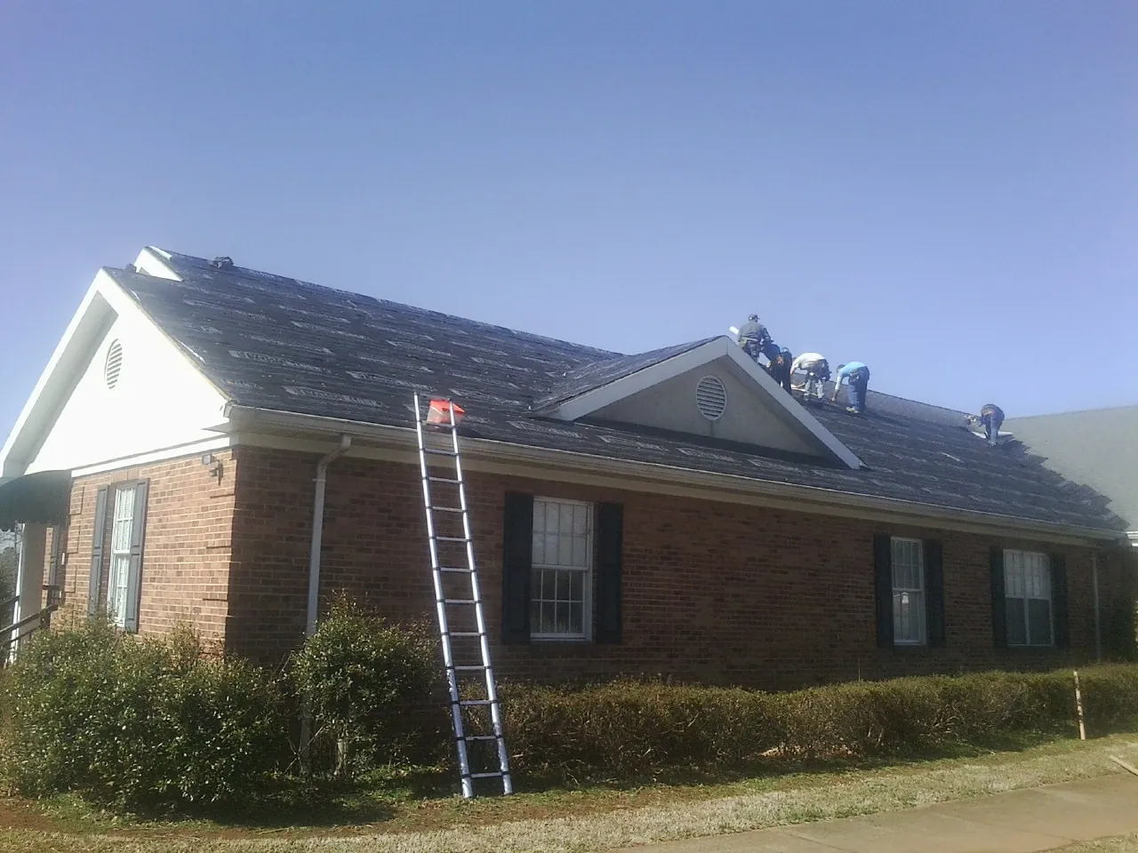 Crew tearing shingles off a brick ranch roof with a ladder set — Emergency Roof Repair work by our crew in Cedar Creek